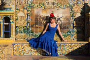 Brunette woman sits on the edge of a fountain in blue flamenco dress under a sign that says Madrid in Spain