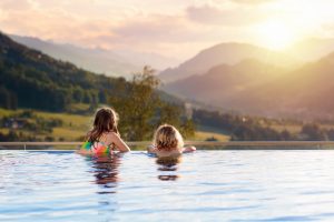 Kids play in outdoor infinity swimming pool of luxury spa alpine resort at sunset in Alps mountains. Spring or summer vacation for family with children. Boy and girl in hot tub with mountain view.